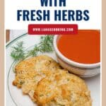 Two drop biscuits with fresh herbs on a plate, served with a bowl of tomato soup and a dill garnish.
