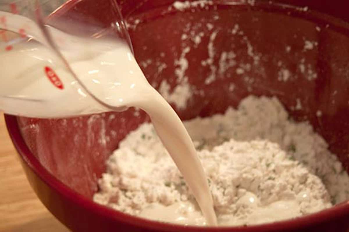 Milk being poured from a measuring cup into a bowl of flour mixture for baking.