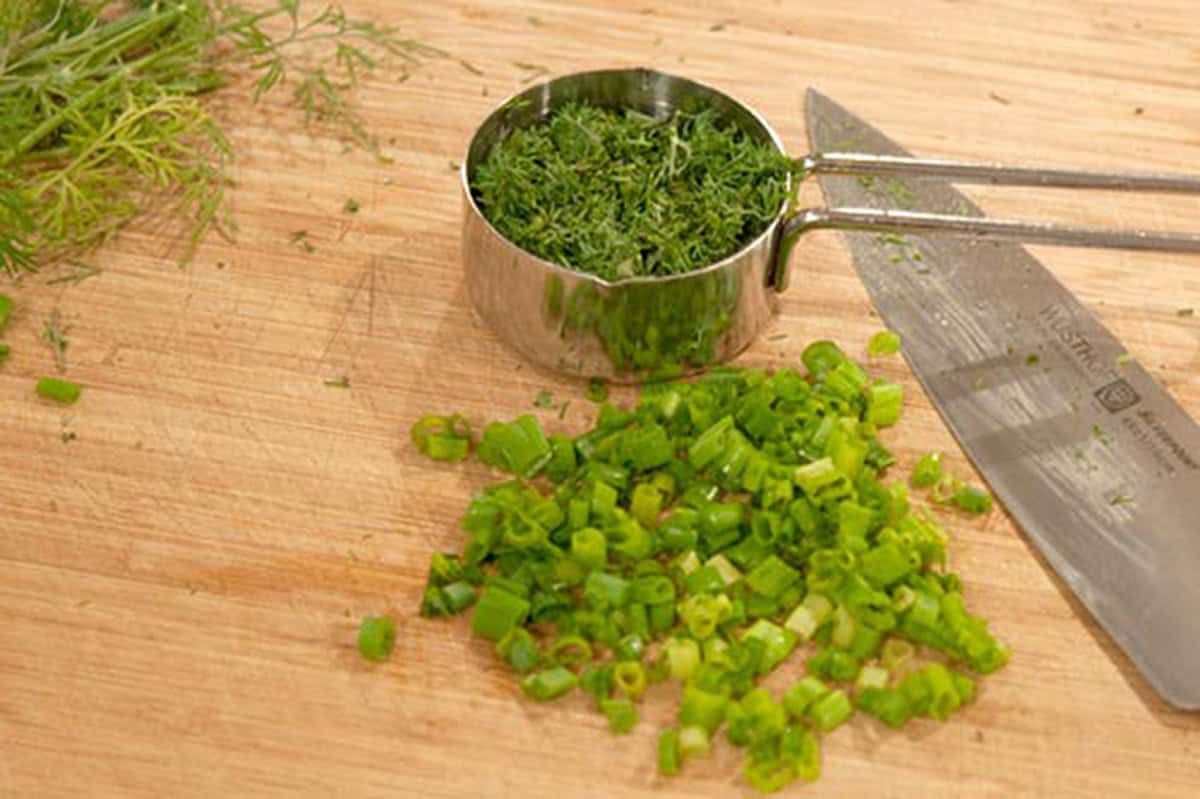Chopped green onions and a metal cup of chopped herbs on a wooden cutting board with a knife.