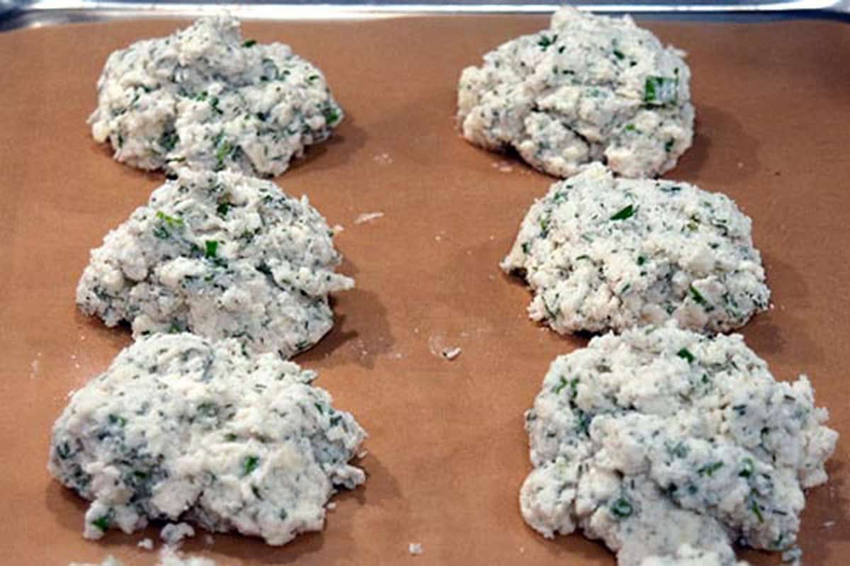 Six unbaked herb biscuit dough mounds on a parchment-lined baking sheet.