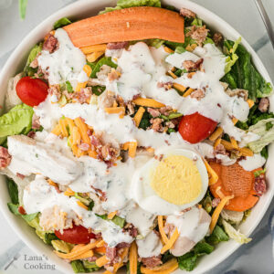 A bowl of salad with lettuce, grape tomatoes, shredded cheddar cheese, bacon bits, sliced carrots, grilled chicken, a boiled egg, and ranch dressing on top.