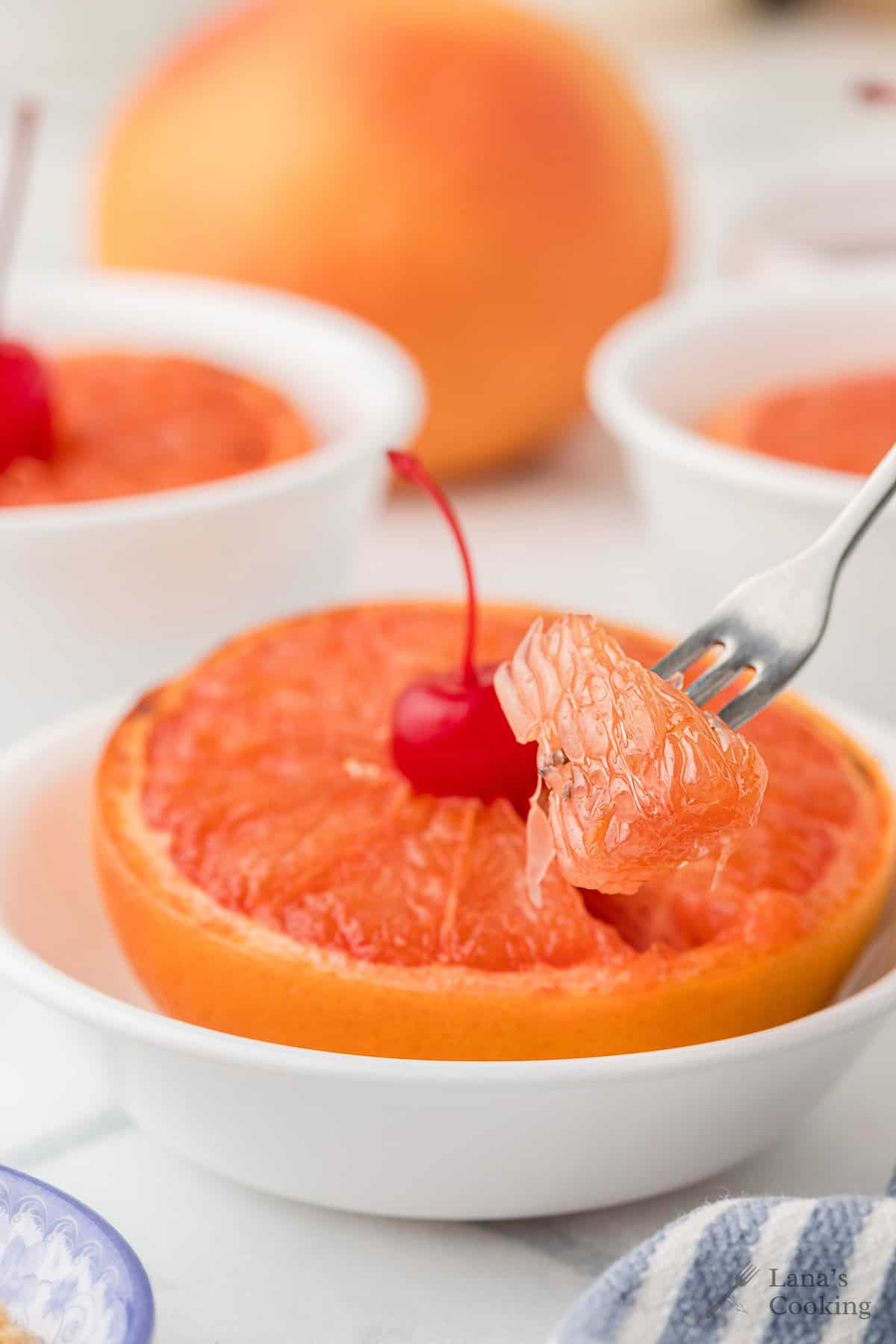 A fork holds a grapefruit segment with a cherry on top, resting in a white bowl with more grapefruit bowls nearby.