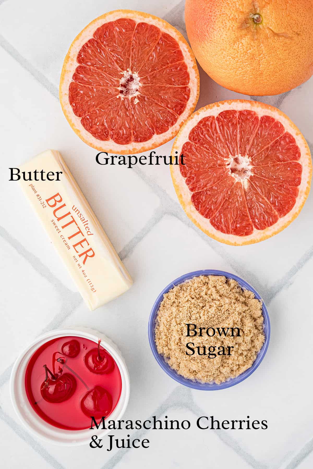 Halved grapefruit, a stick of butter, a bowl of brown sugar, and maraschino cherries with juice on a white surface.