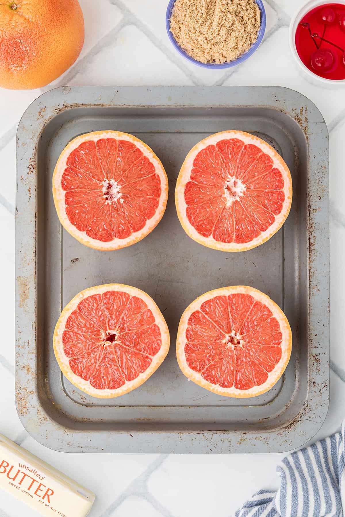 Four grapefruit halves on a baking sheet, with brown sugar, cherries, and butter nearby on a marble surface.