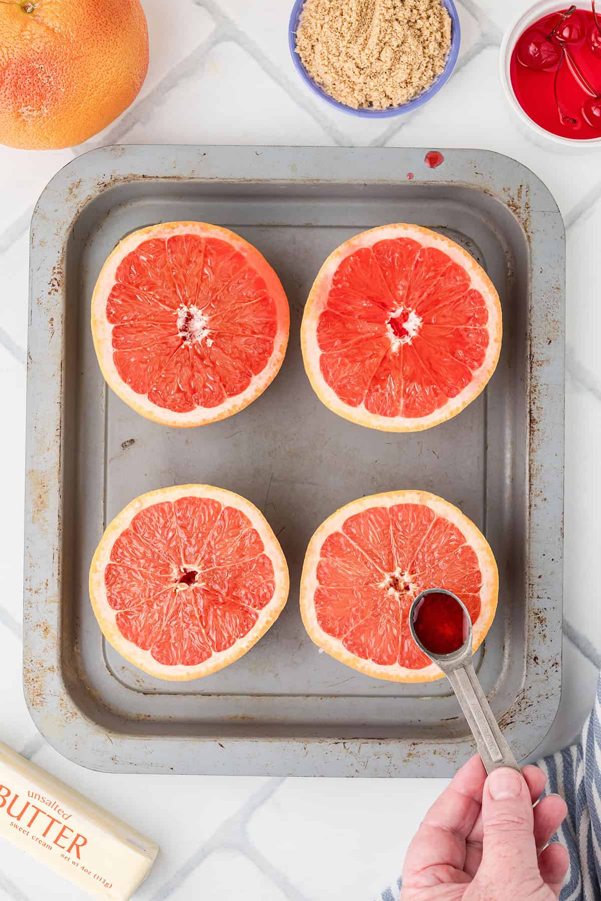 Four grapefruit halves on a baking sheet; a hand pours cherry juice on one. Brown sugar and cherries nearby.