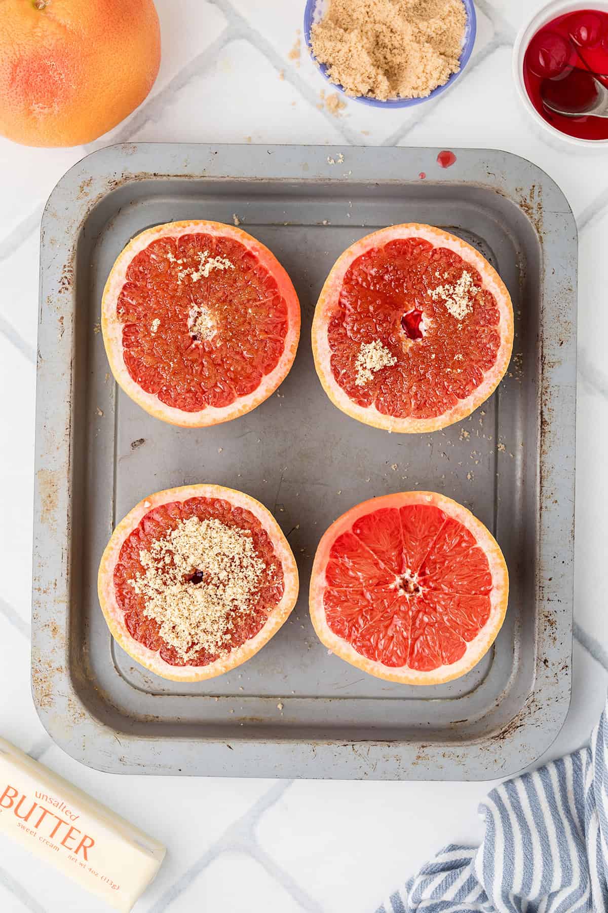 Four grapefruit halves on a baking tray, some topped with brown sugar; butter and toppings nearby.