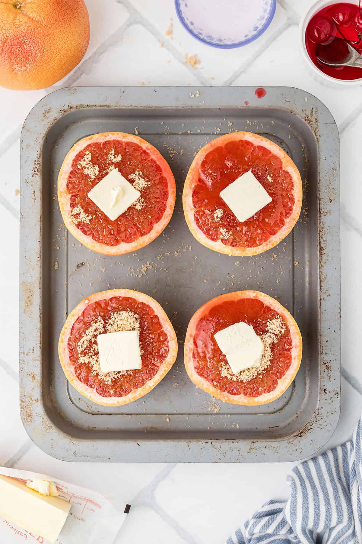 Four grapefruit halves on a baking tray, each topped with brown sugar and a pat of butter, ready to bake.