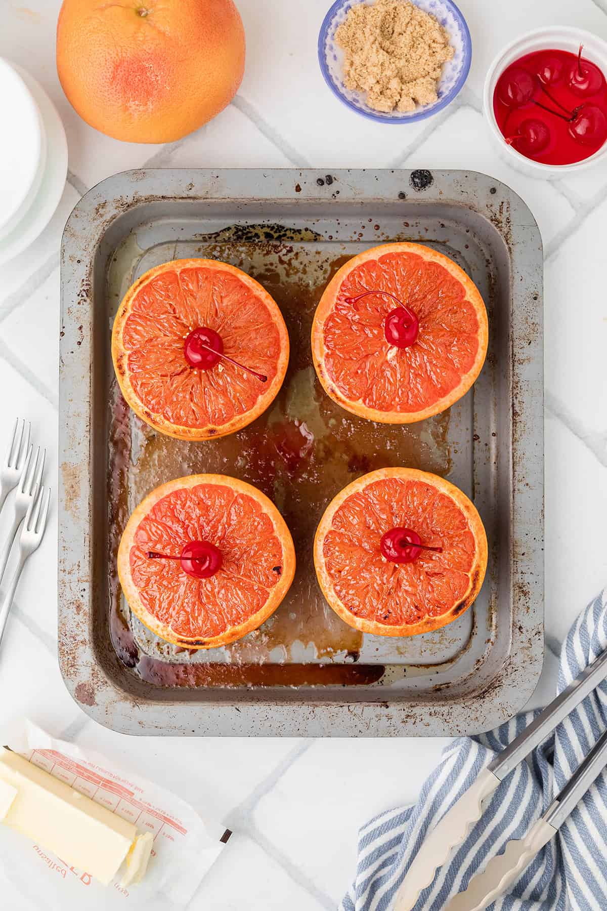 Four halved grapefruits with cherries on top in a baking tray, surrounded by ingredients on a white surface.