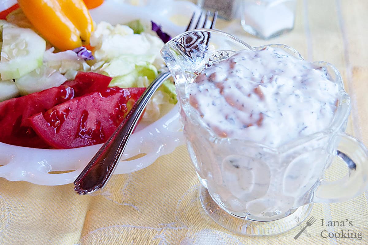 A glass pitcher of creamy salad dressing sits next to a plate of fresh salad with tomatoes, cucumbers, lettuce, and peppers on a yellow tablecloth.