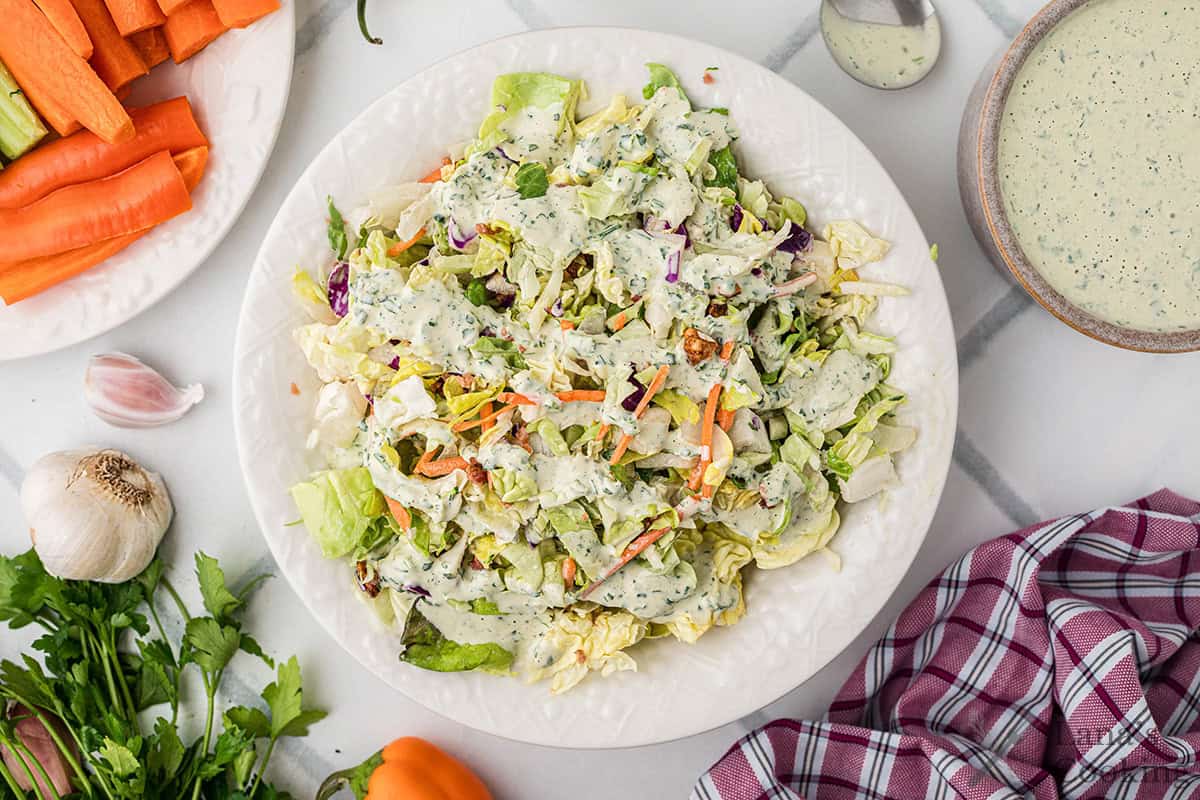 Chopped salad with creamy herb dressing on a white plate, surrounded by vegetables and a red plaid napkin.