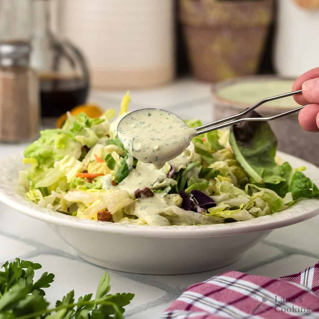 A spoon drizzles creamy dressing over a fresh green salad in a white bowl on a kitchen counter.