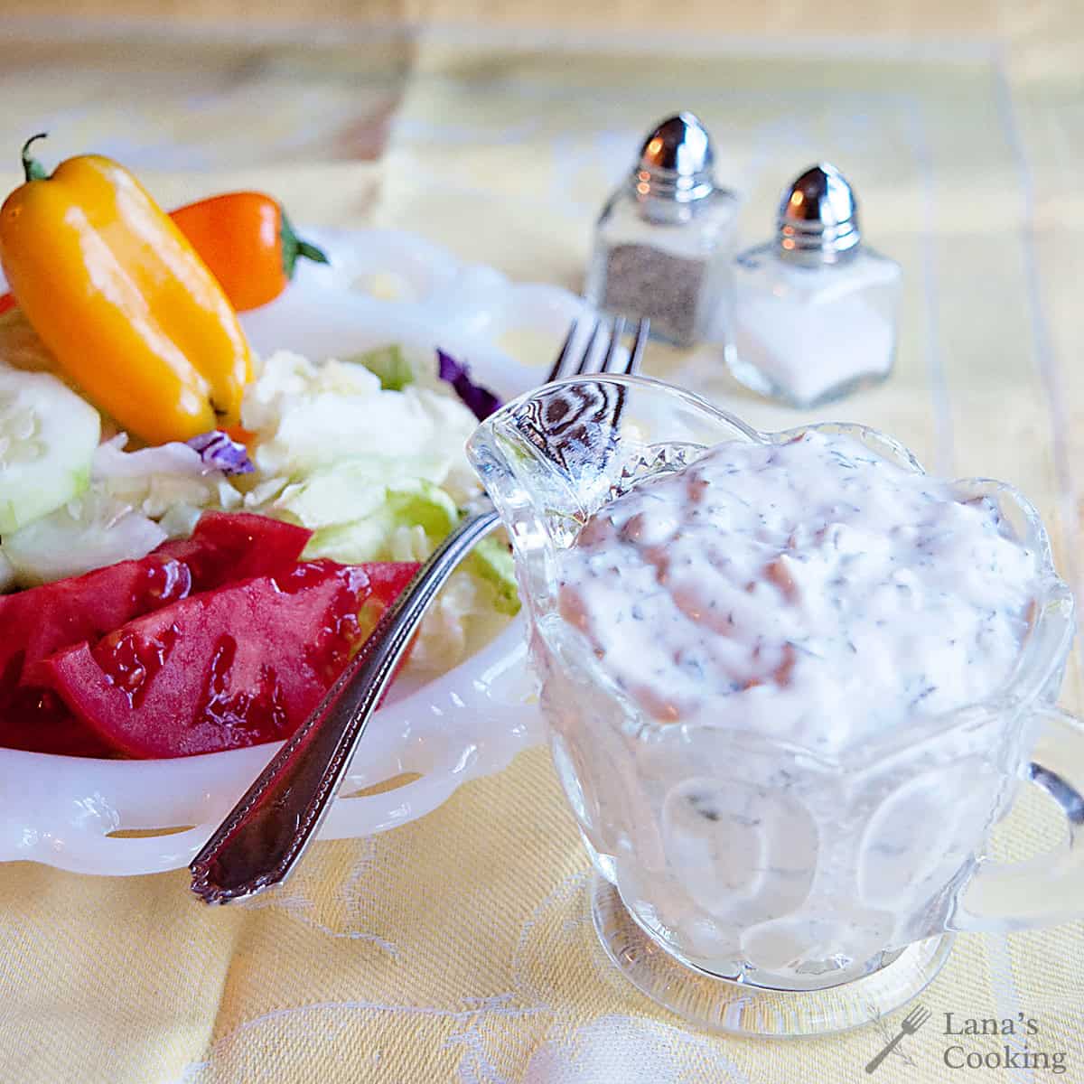 A glass cup of creamy salad dressing sits next to a plate of fresh lettuce, tomato, and mini bell peppers on a yellow tablecloth with salt and pepper shakers in the background.