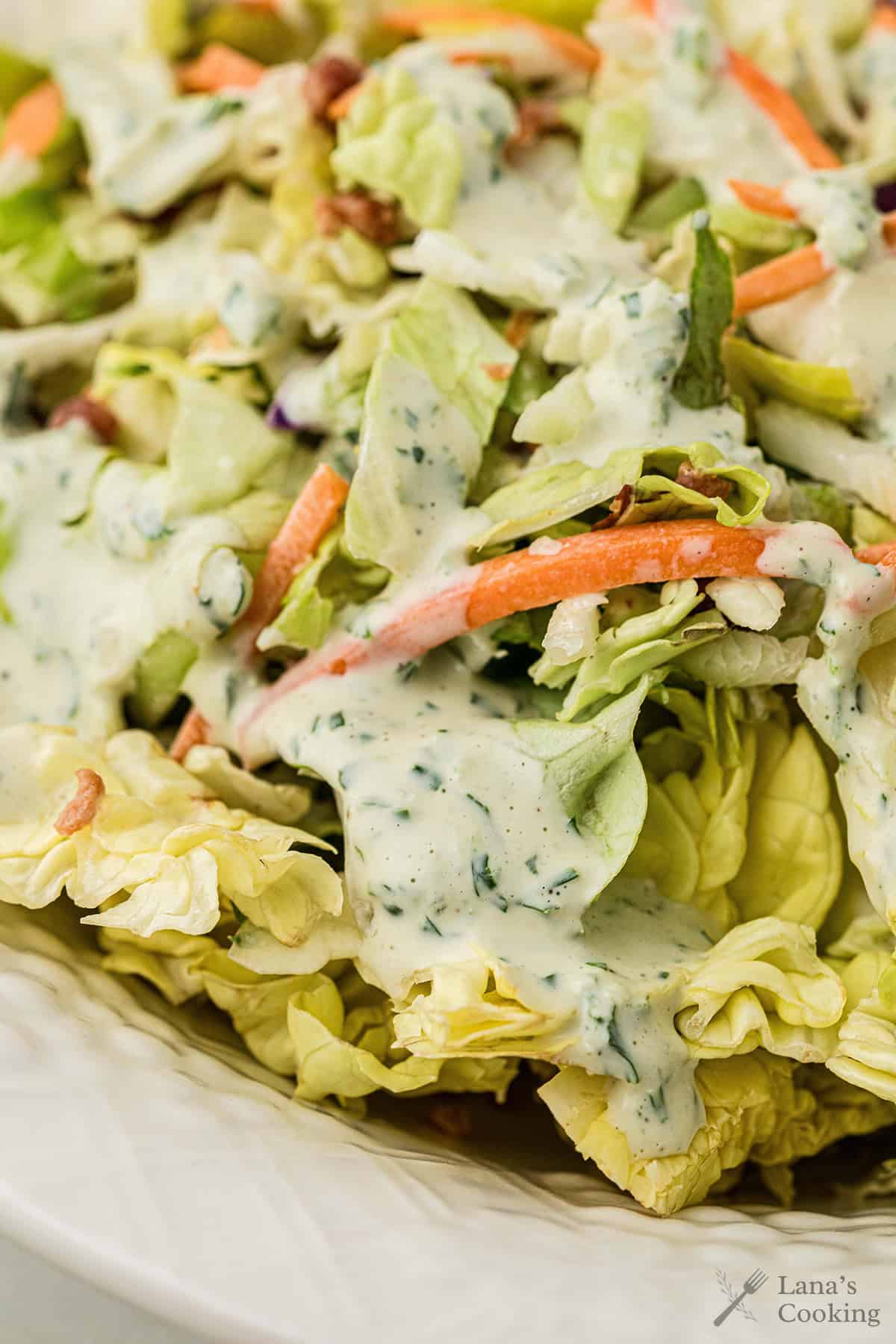 Close-up of a fresh salad with lettuce, carrots, and creamy ranch dressing on top in a white bowl.