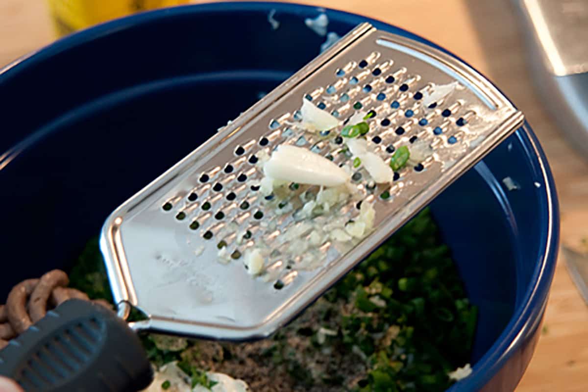 A metal grater with chopped garlic and green herbs resting on top of a blue bowl filled with chopped ingredients.