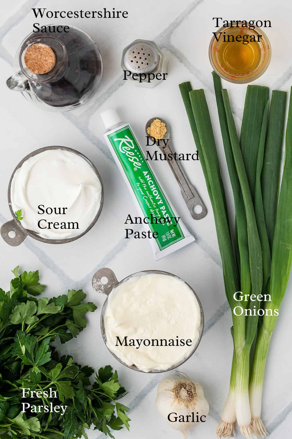 Ingredients for a dressing, including mayonnaise, sour cream, anchovy paste, herbs, and seasonings on a white surface.