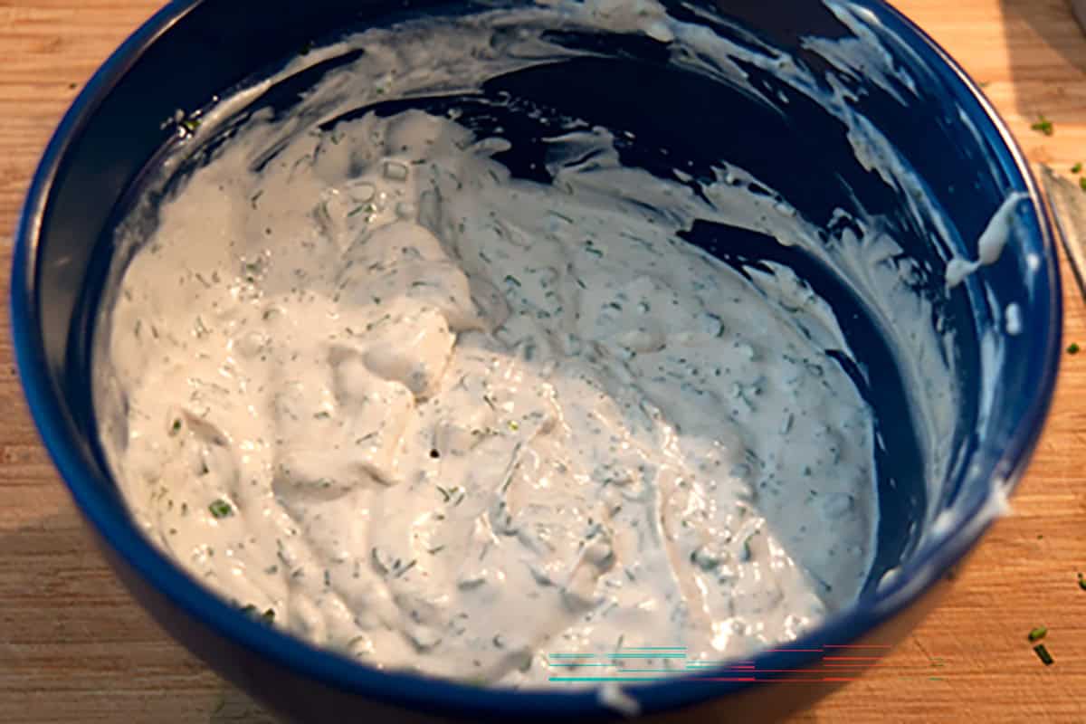 A blue bowl containing a thick, white, herb-speckled mixture or dip sits on a wooden surface.