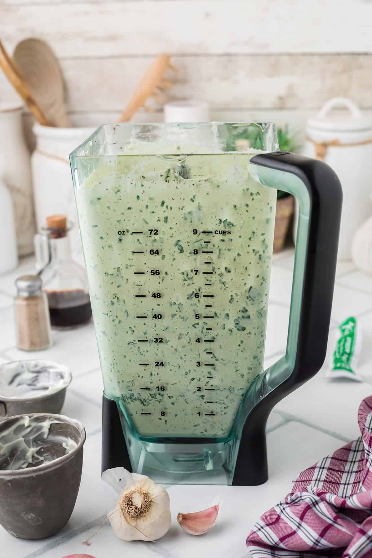 A blender filled with creamy green goddess dressing sits on a kitchen counter with ingredients and utensils nearby.