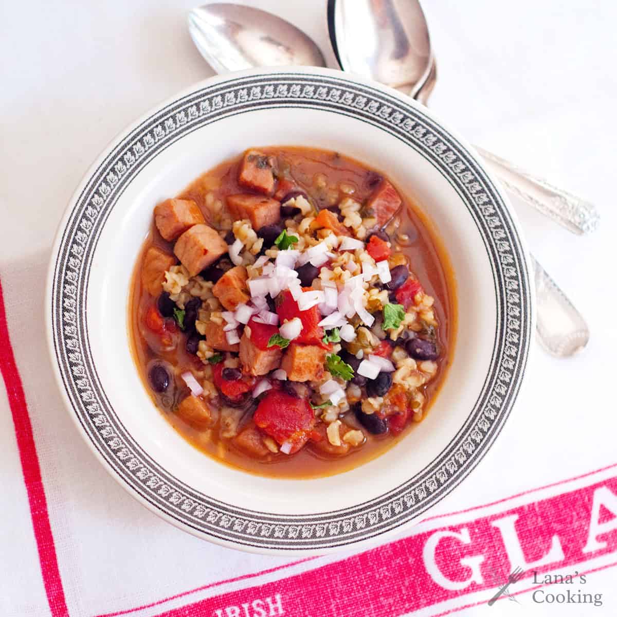 A bowl of soup with beans, diced sausage, rice, and vegetables sits on a patterned plate; two spoons rest above the bowl on a white cloth with red text and a visible “Lana’s Cooking” watermark.