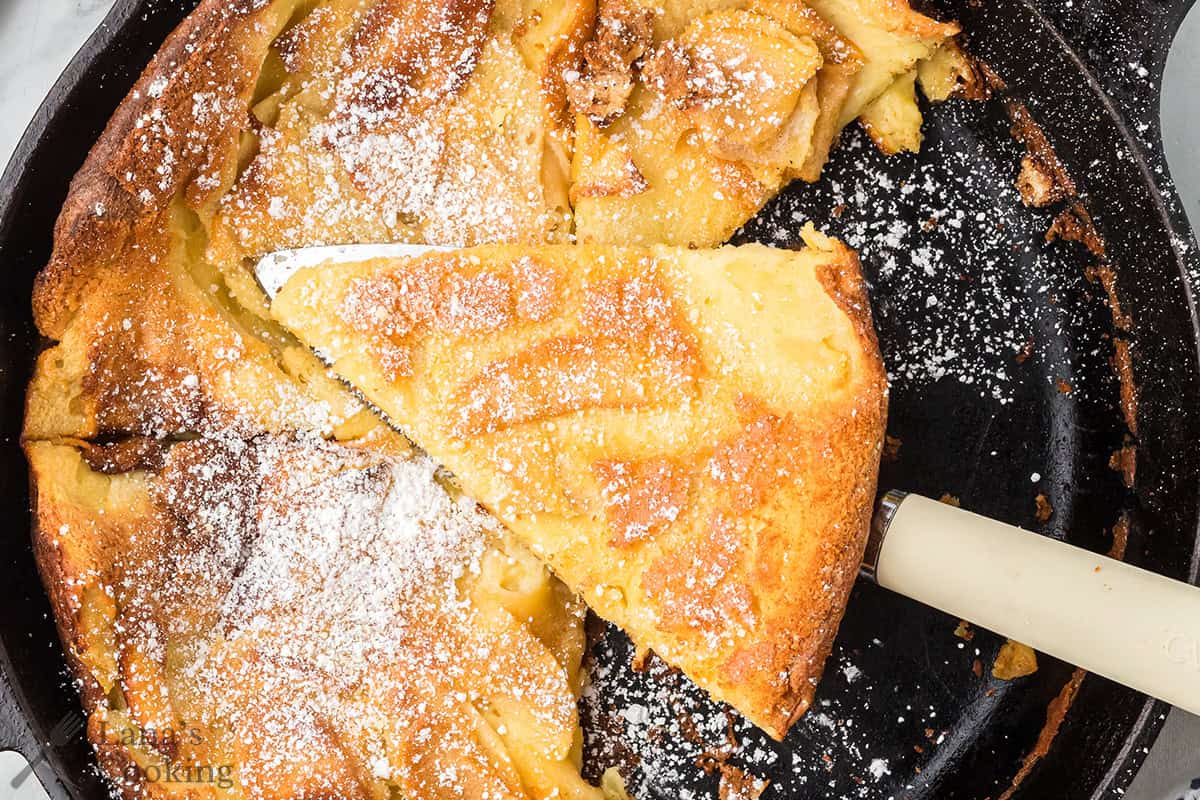 A slice of baked pancake dusted with powdered sugar in a skillet, with a spatula lifting a piece.