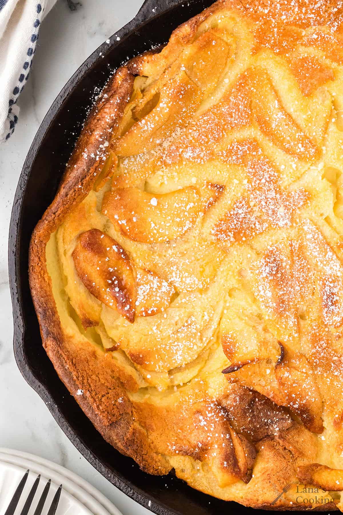 Golden Dutch baby pancake in a skillet, topped with powdered sugar, with visible baked apple slices.