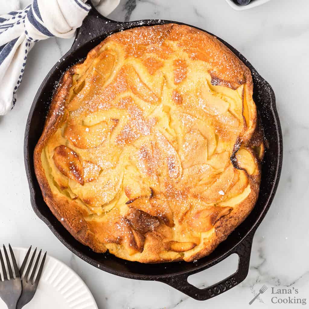 A golden-brown apple Dutch baby pancake in a cast iron skillet, dusted with powdered sugar.