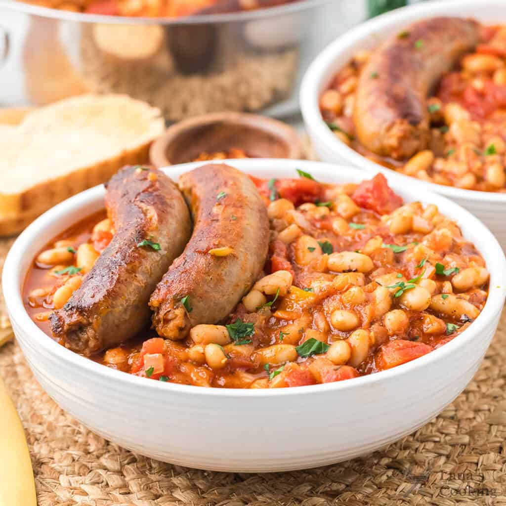 A bowl of white beans topped with two sausages, with bread and another bowl in the background.