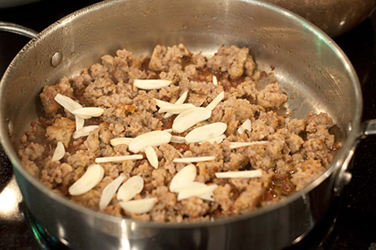 Ground sausage and sliced garlic cooking in a stainless steel skillet on a stove.