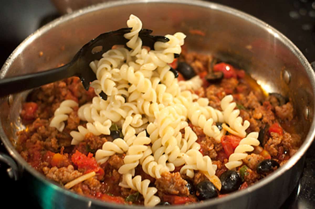 A pot of rotini pasta with ground meat, tomatoes, and black olives being mixed with a slotted spoon.