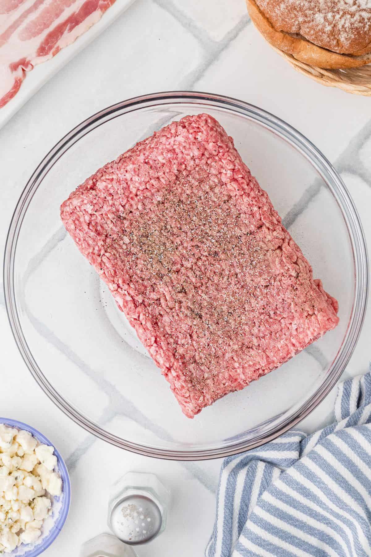 Ground beef in a glass bowl with seasonings added, but not mixed in.