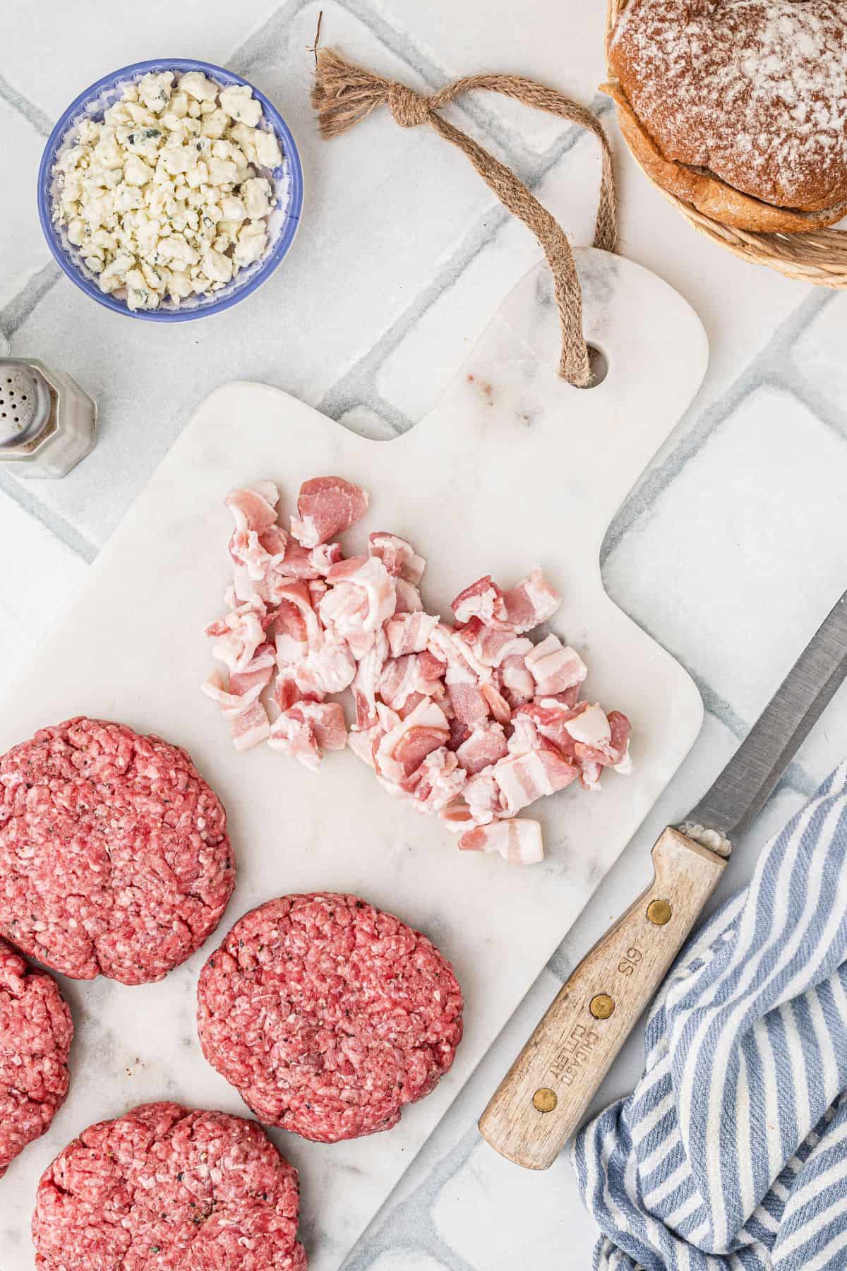 Bacon chopped on a white cutting board pictured next to four uncooked hamburger patties. 