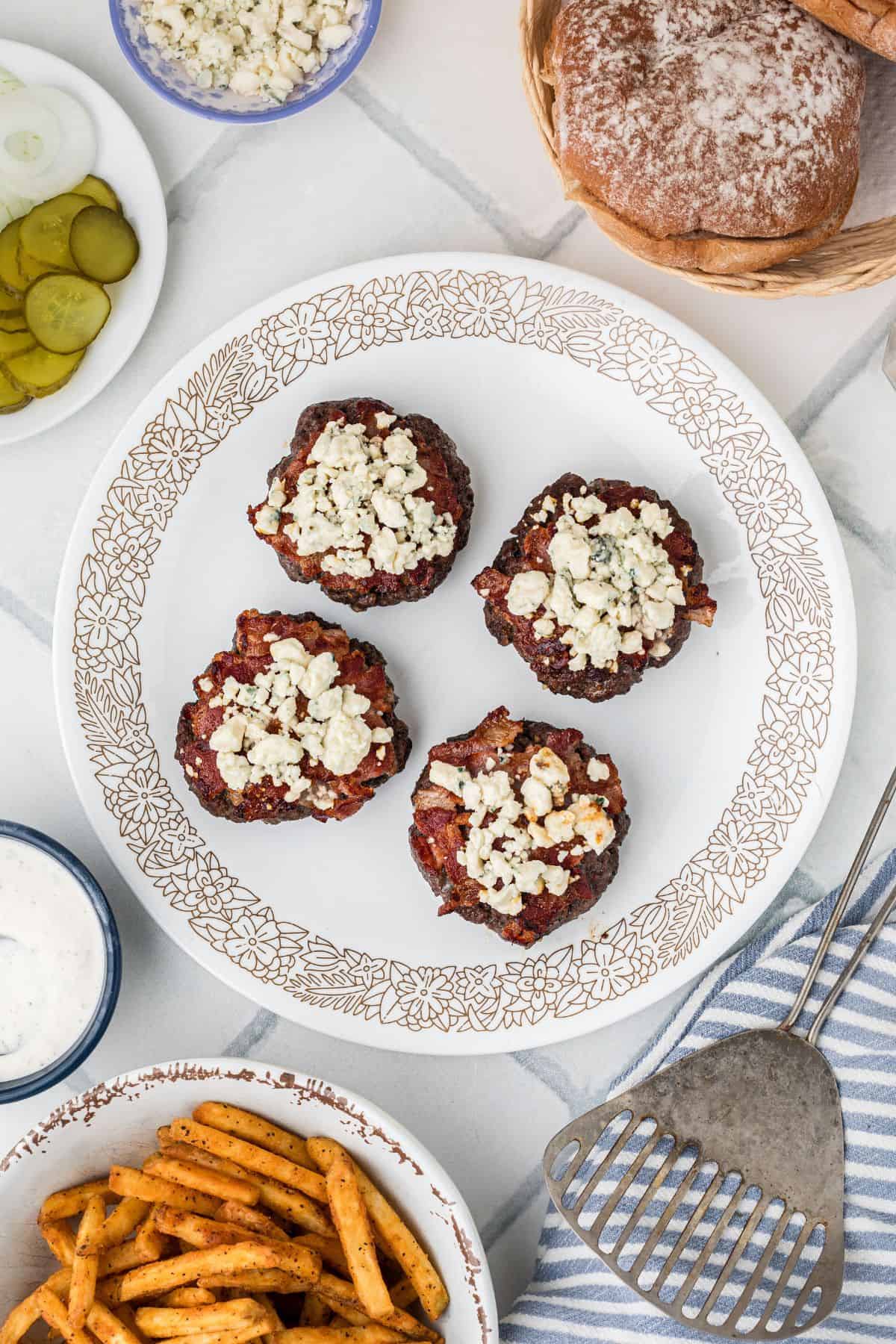Blue cheese crumbles on top of cooked burger patties on a white plate with a brown floral patters on the outer edge of the plate.
