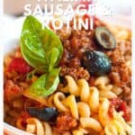 A bowl of rotini pasta with Italian sausage, tomato sauce, olives, and a fresh basil leaf on top.