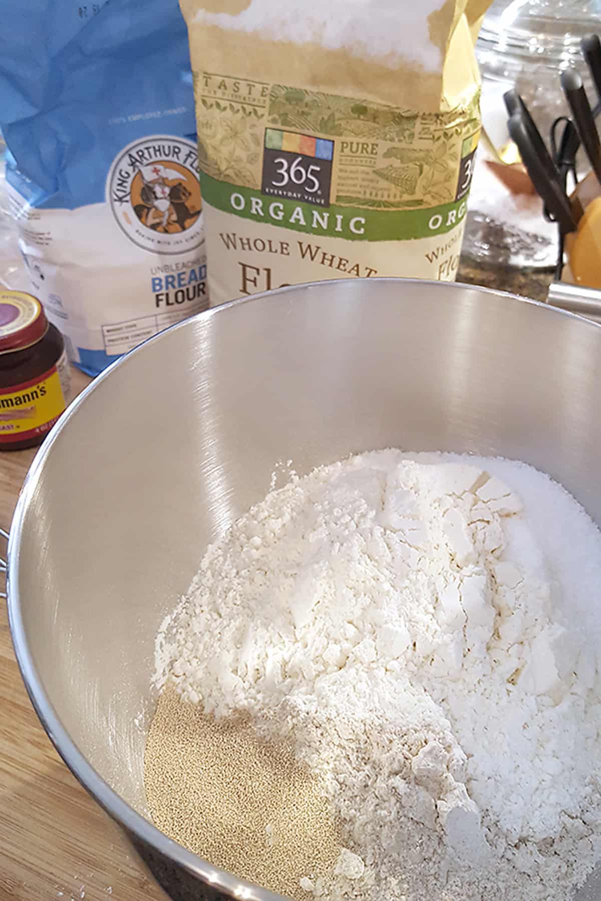 A mixing bowl containing flour and yeast, with bags of bread and whole wheat flour in the background.