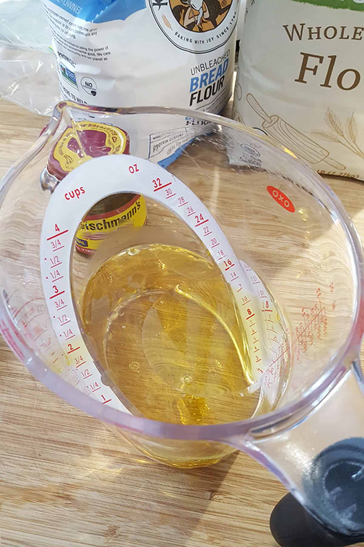 A clear plastic measuring cup containing oil, with bags of flour and a jar of yeast in the background.