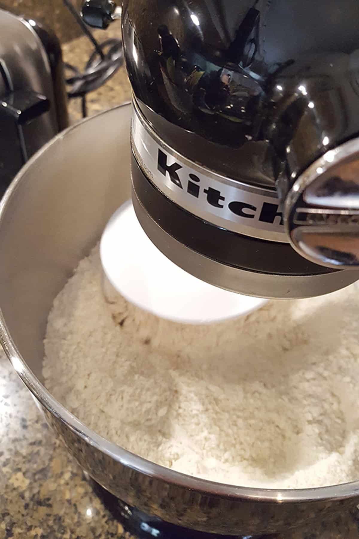 KitchenAid stand mixer with a dough hook mixing flour in a metal bowl.