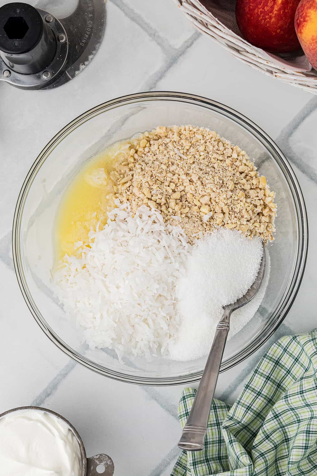 A glass bowl containing shredded coconut, chopped nuts, sugar, and melted butter, with a spoon resting inside. Nearby are peaches, a food processor, and a green checkered cloth.