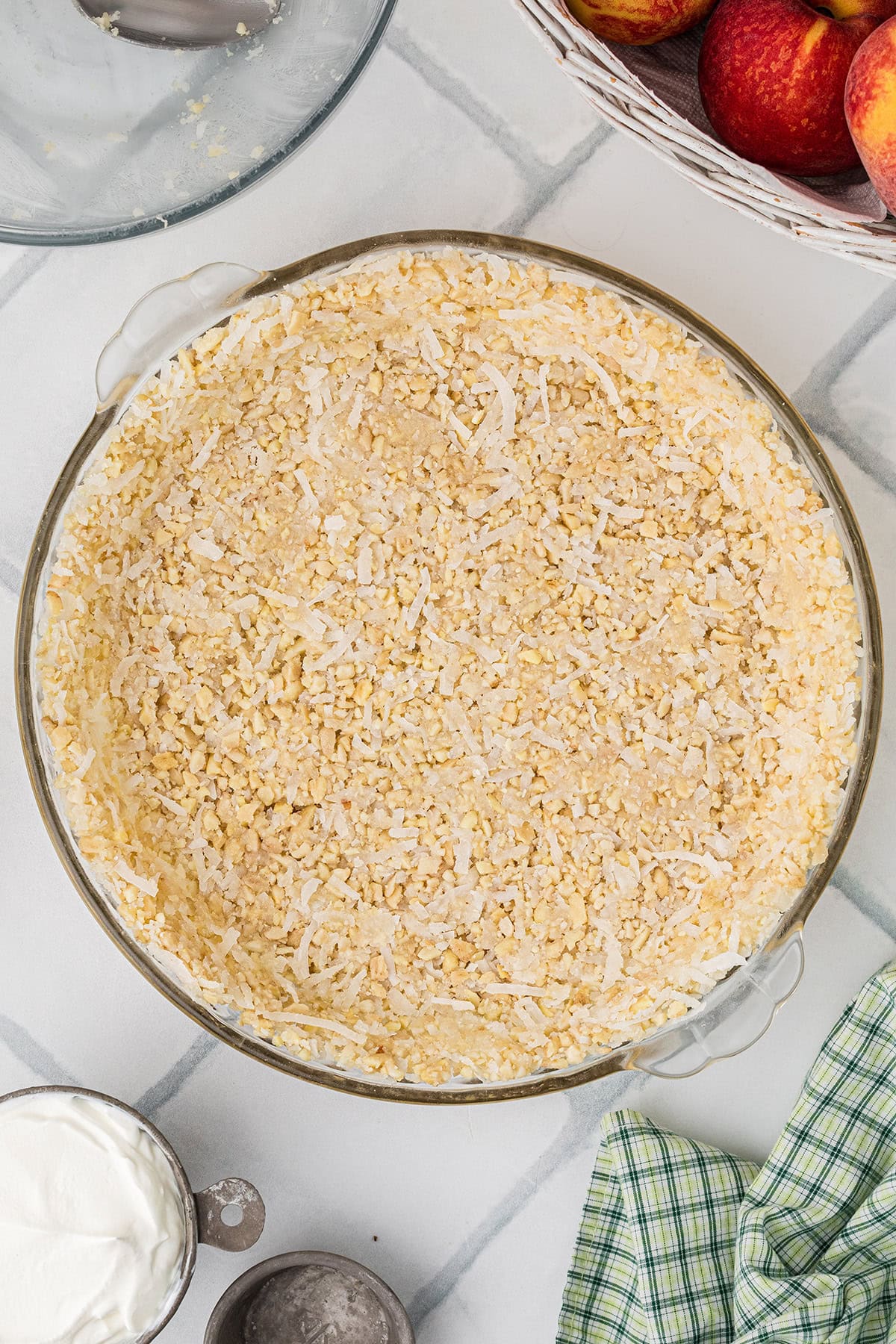 A glass pie dish filled with an unbaked crumbly crust mixture, next to a bowl of peaches, a green checkered towel, and a bowl of white cream.
