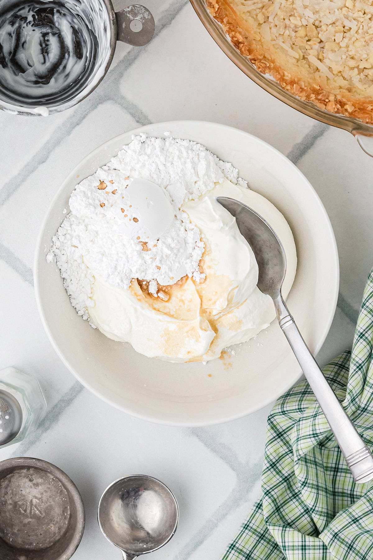 A white bowl containing powdered sugar, cream cheese, sour cream, and vanilla, with a spoon inside. Baking tools and a pie crust are nearby on a white surface.