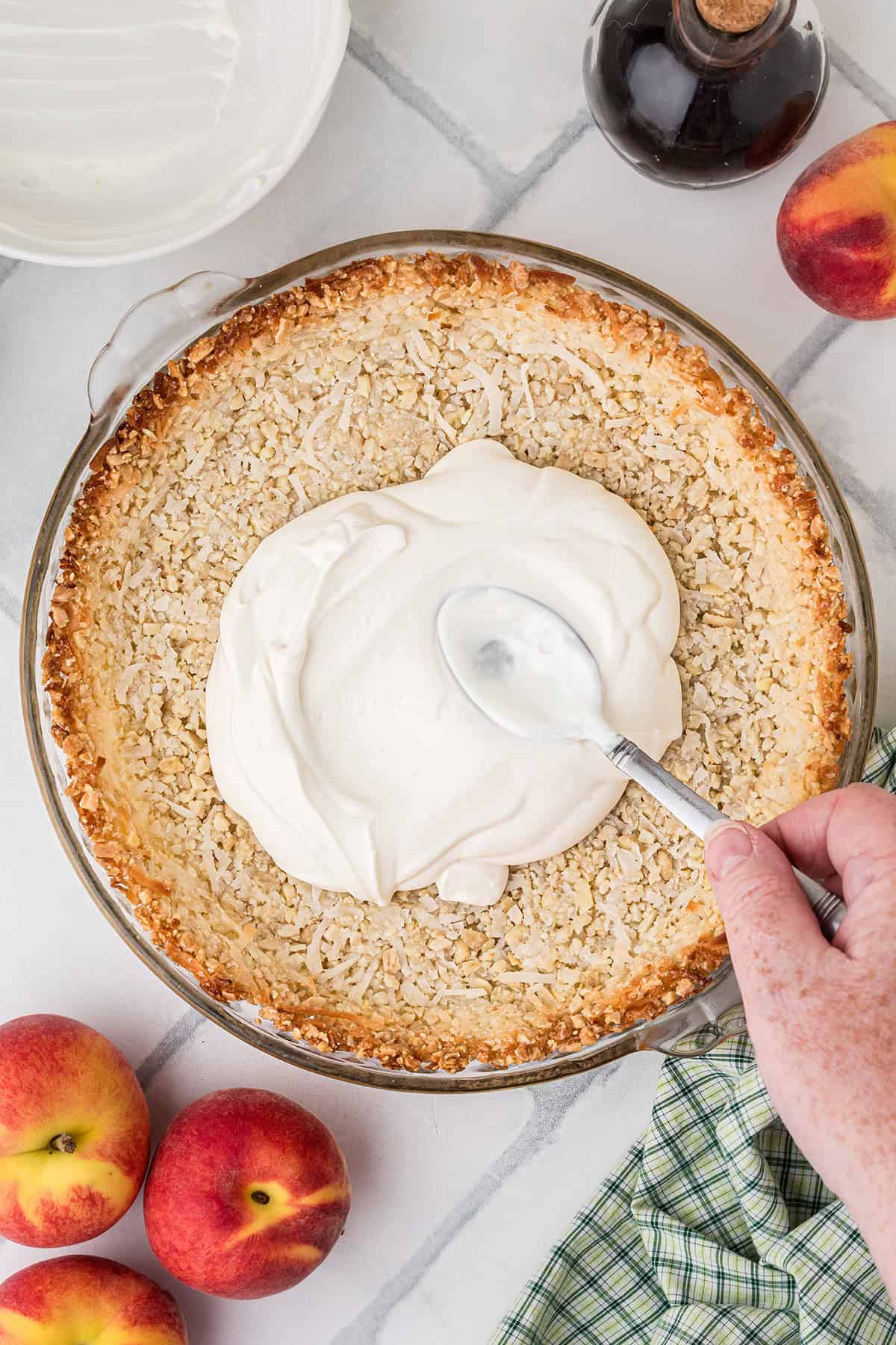 A hand spreads creamy filling over a baked pie crust with a spoon. Fresh peaches, a glass bottle, and a checkered cloth are nearby on a white surface.
