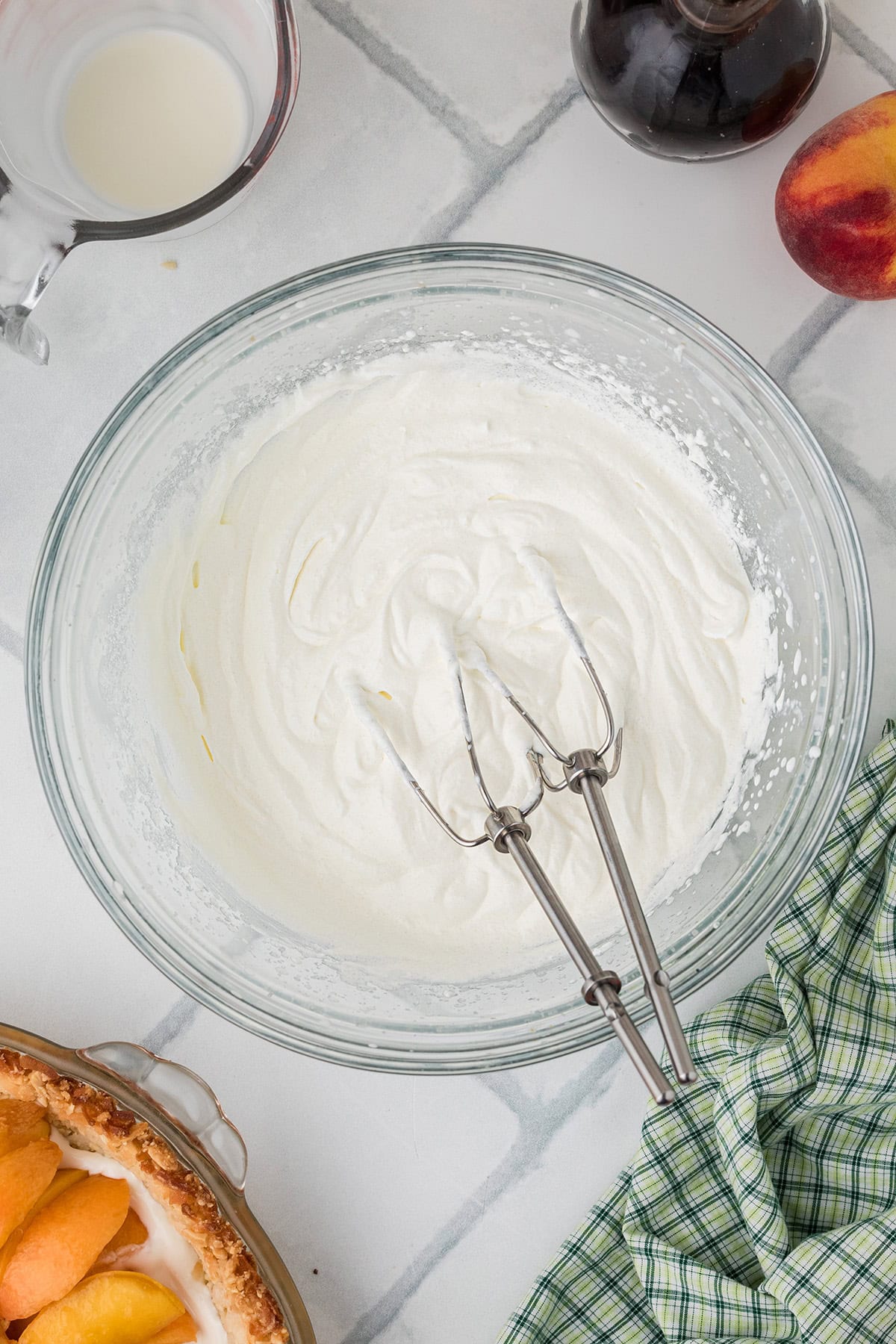 A glass bowl with whipped cream and mixer beaters on a tiled countertop, surrounded by a glass of milk, a peach, syrup bottle, and part of a peach pie.