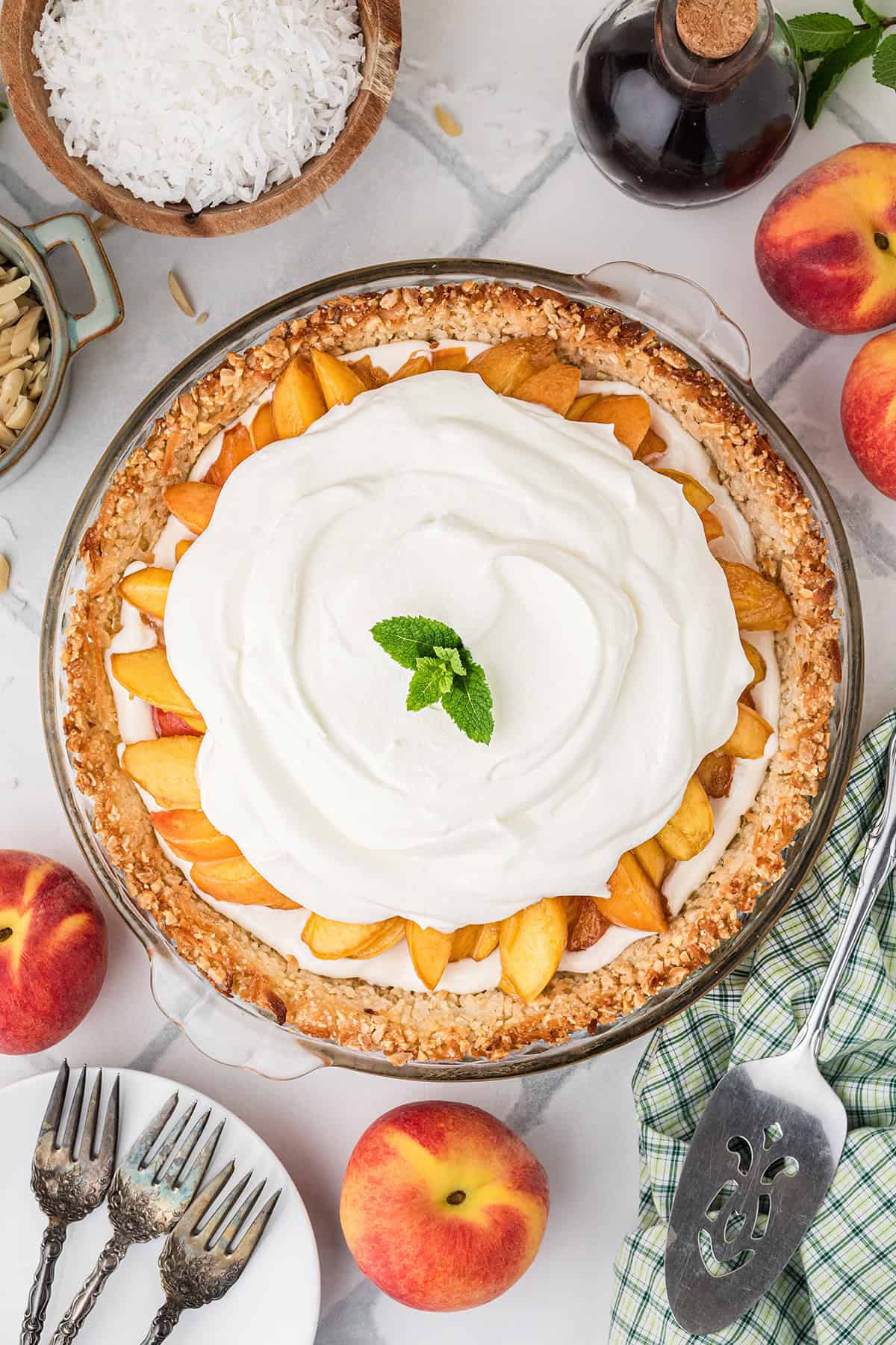 Peach pie topped with whipped cream and a mint leaf, surrounded by whole peaches, a pie server, forks, shredded coconut, and a bottle of vanilla extract.