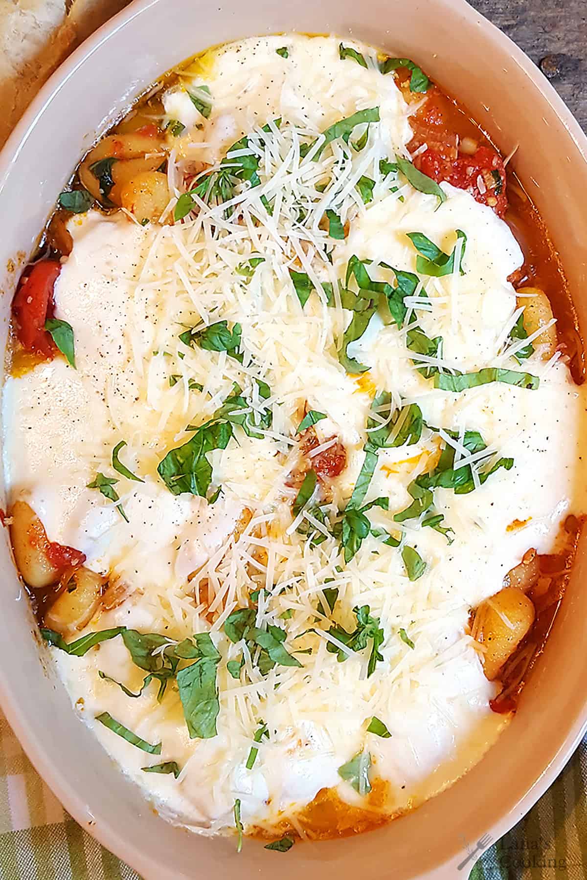 A baked dish of gnocchi topped with melted cheese, tomato sauce, grated parmesan, and chopped fresh basil in an oval ceramic baking dish.