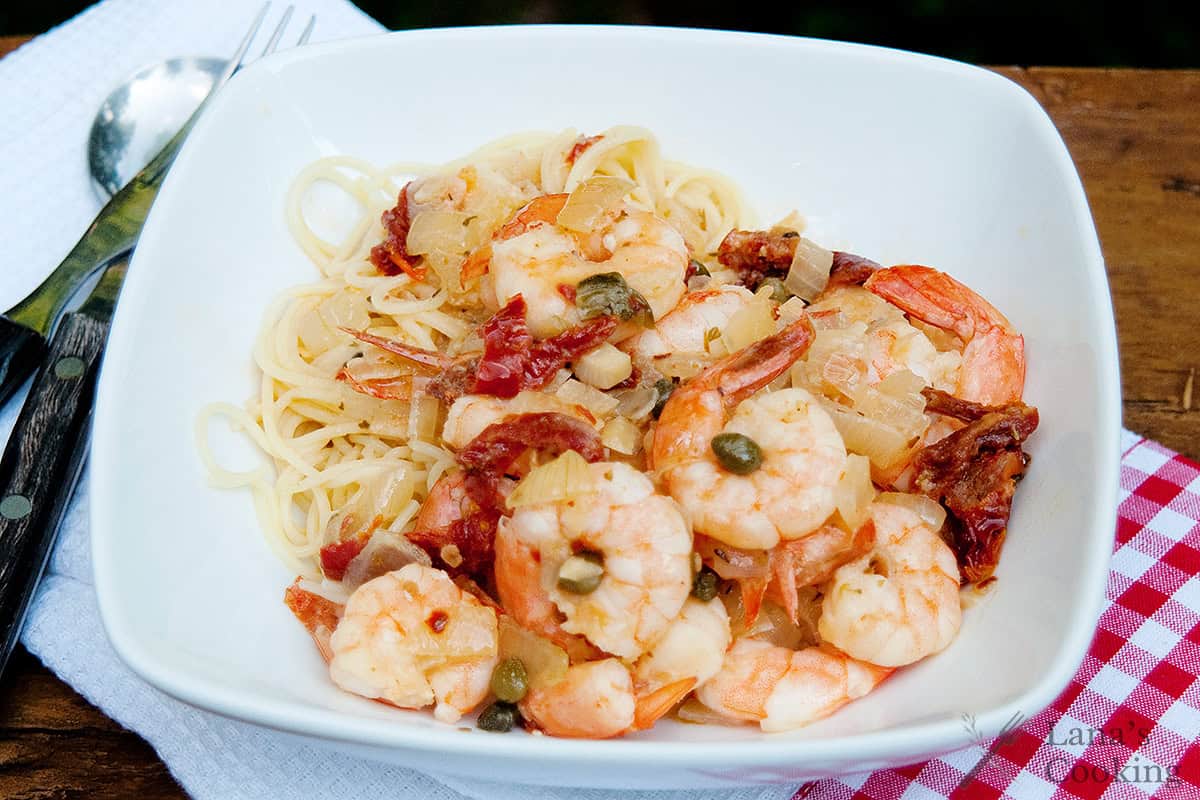 A bowl of pasta topped with shrimp, sun-dried tomatoes, capers, and onions, served on a wooden table with a fork and spoon nearby.
