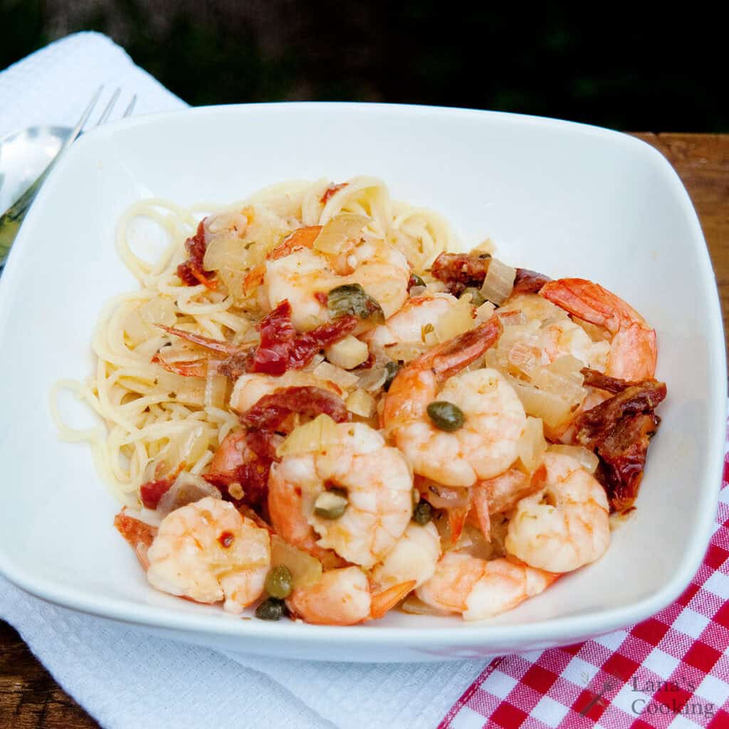 A white bowl of angel hair pasta topped with cooked shrimp, capers, diced onions, and sun-dried tomatoes, placed on a white napkin with a red checkered edge.