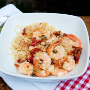 A white bowl of angel hair pasta topped with cooked shrimp, capers, diced onions, and sun-dried tomatoes, placed on a white napkin with a red checkered edge.