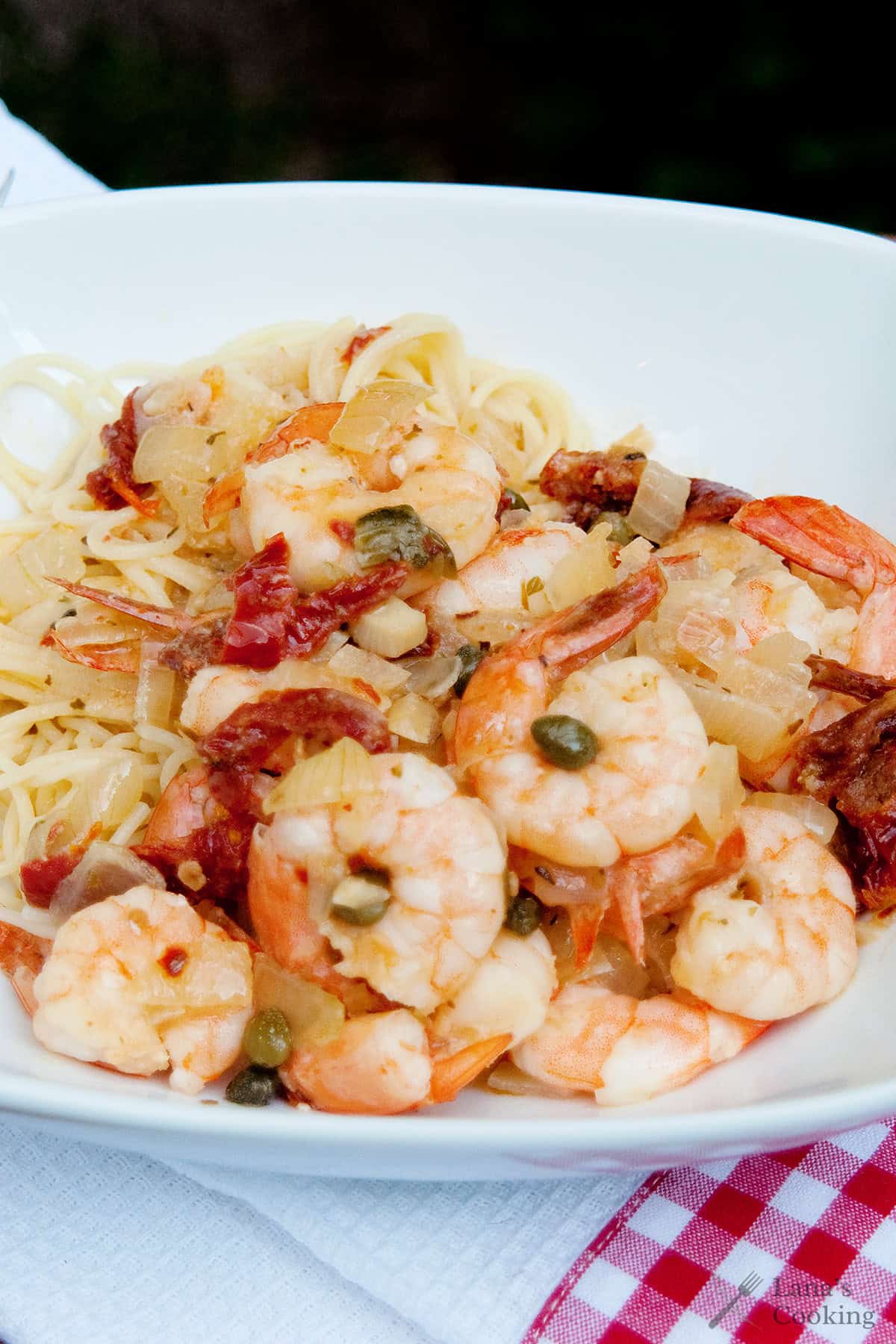 A plate of pasta topped with shrimp, capers, onions, and sun-dried tomatoes, served on a white dish with a red and white checkered napkin underneath.