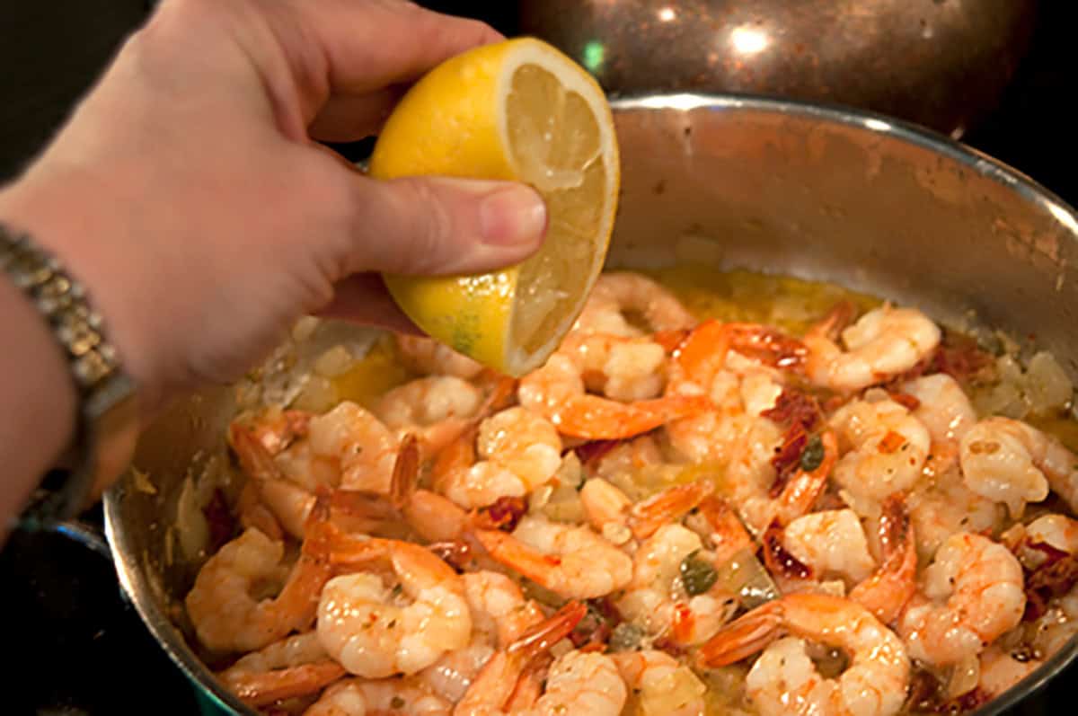 A hand squeezes a lemon half over a pan of cooked shrimp with sauce.