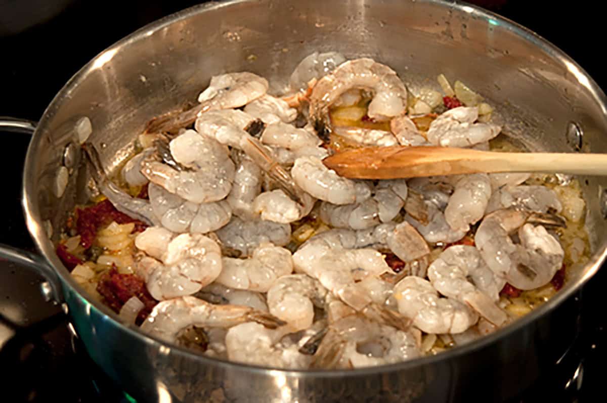 Raw shrimp being sautéed with chopped onions and other ingredients in a stainless steel pan with a wooden spoon.