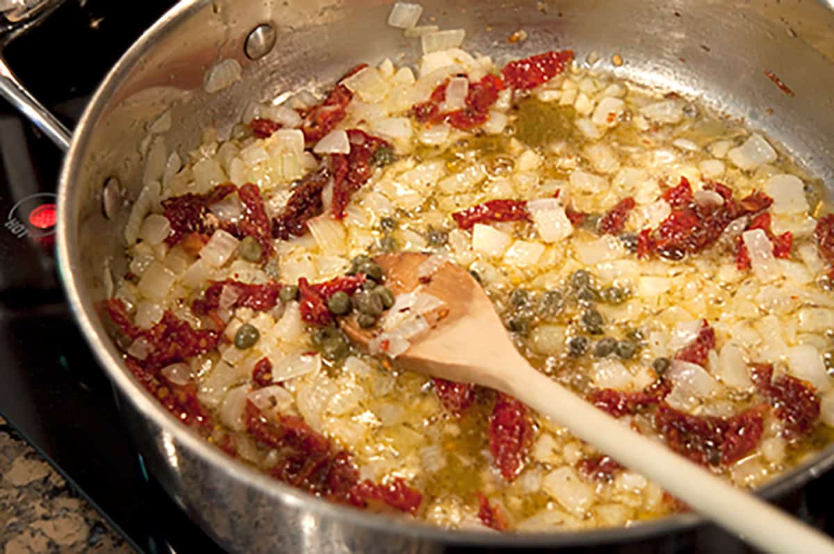 Chopped onions, sun-dried tomatoes, and capers are sautéing in oil in a stainless steel pan, stirred with a wooden spoon.