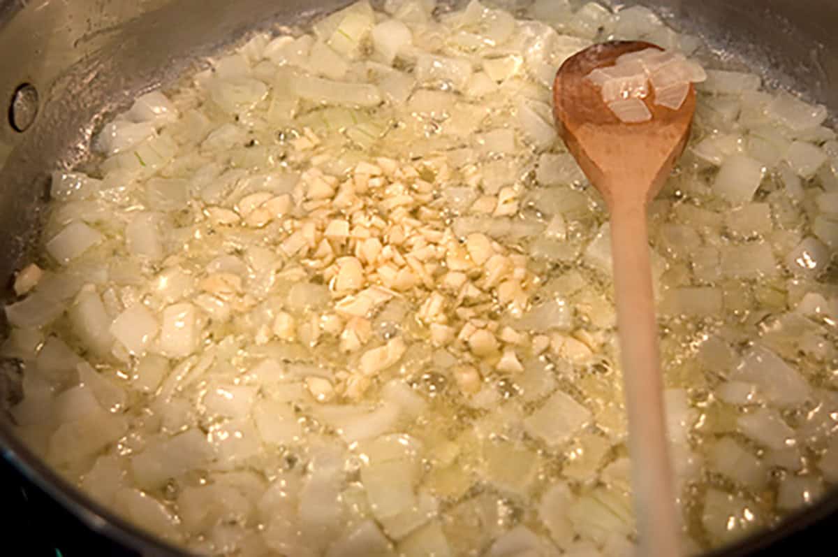 Chopped onions and garlic are sautéing in a pan with butter and oil, being stirred with a wooden spoon.